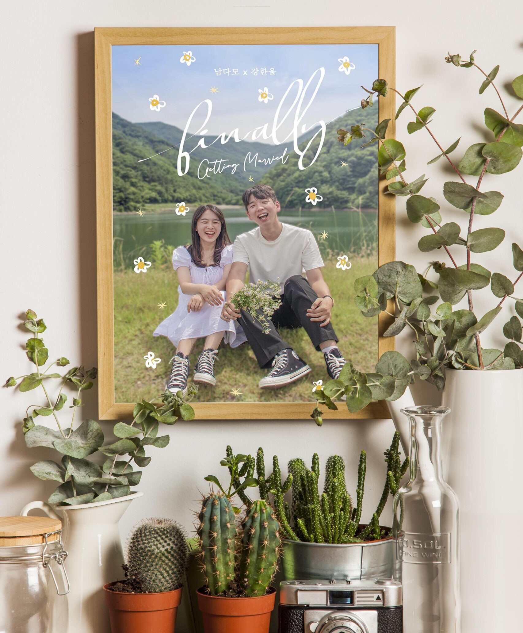 A couple is sitting on the ground in a frame, and the frame is hanging on the wall. There are a lot of plants surrounding the frame.