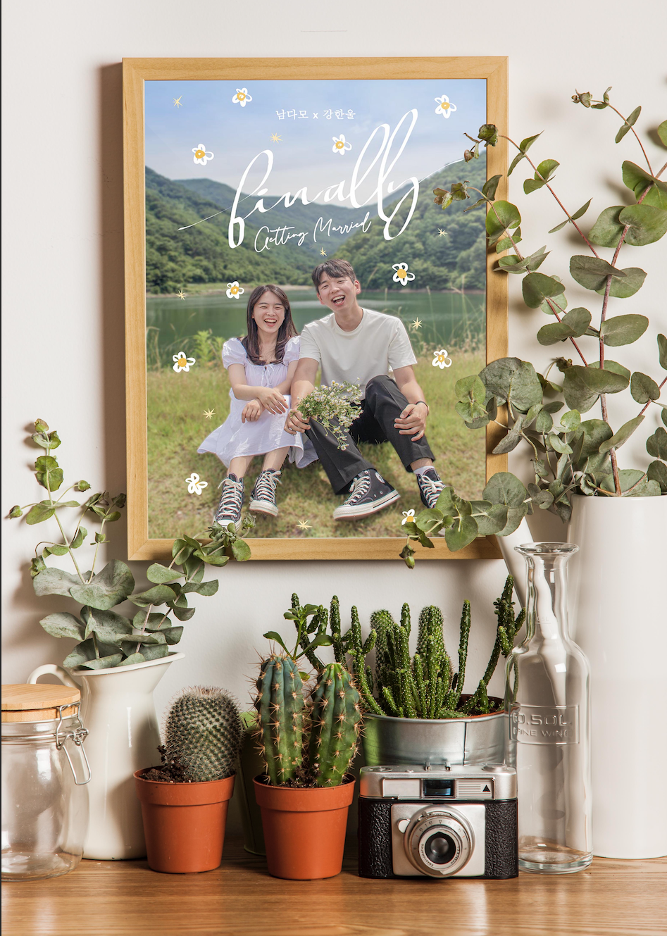 A couple is sitting on the ground in a frame, and the frame is hanging on the wall. There are a lot of plants surrounding the frame.