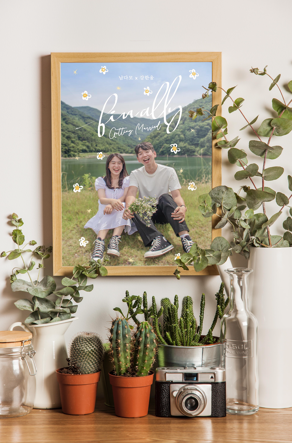 A couple is sitting on the ground in a frame, and the frame is hanging on the wall. There are a lot of plants surrounding the frame.